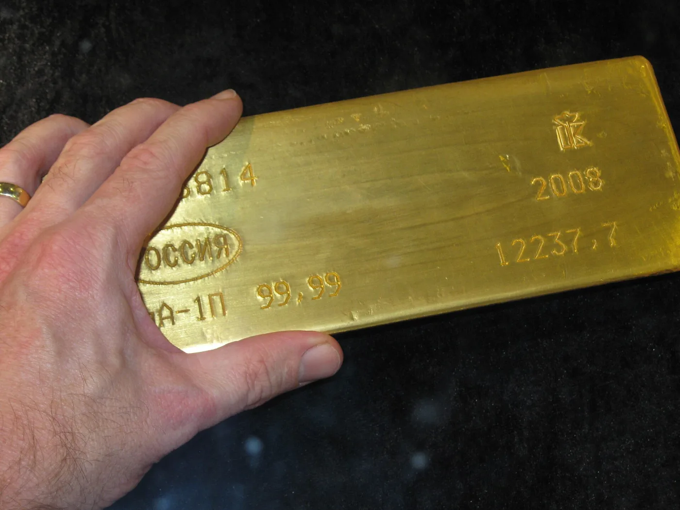 A hand resting on a large gold bar stamped with an oval РОССИЯ mark, 99,99, 2008, 12237,7 and a partly hidden serial number