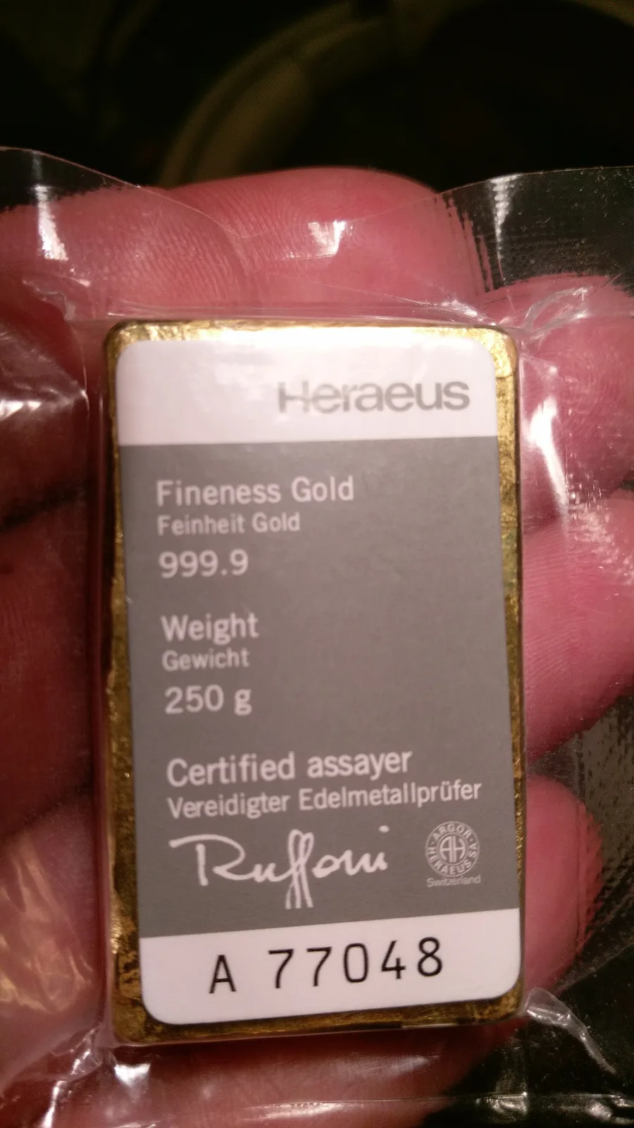 A gold bar in a clear plastic sleeve held in a hand, with a printed Heraeus card reading Fineness Gold 999.9, Weight 250 g, a certified assayer's signature and the serial A 77048
