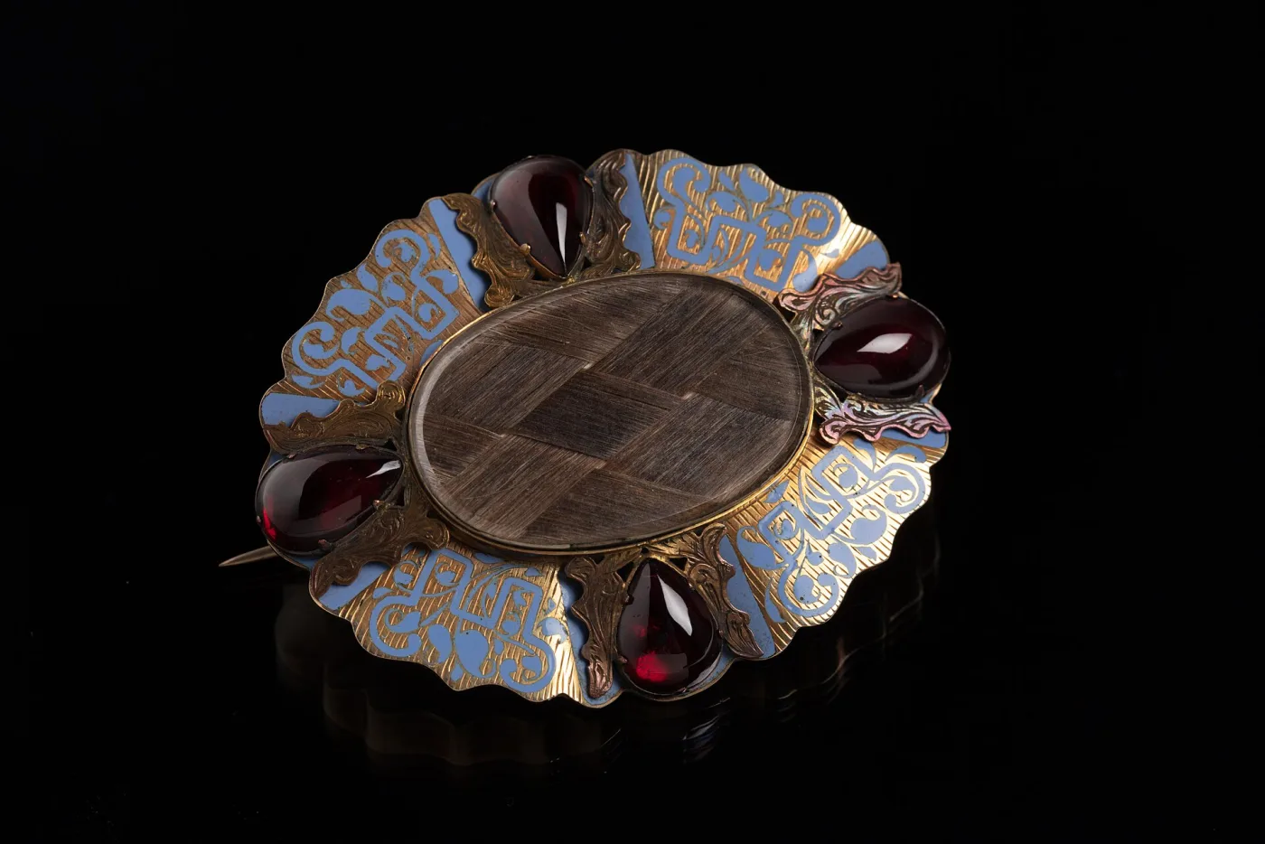 Victorian mourning brooch photographed at an angle on black, with a panel of woven hair under glass, four garnet cabochons and pale blue enamel scrollwork, the point of its pin stem projecting well past the left edge of the mount