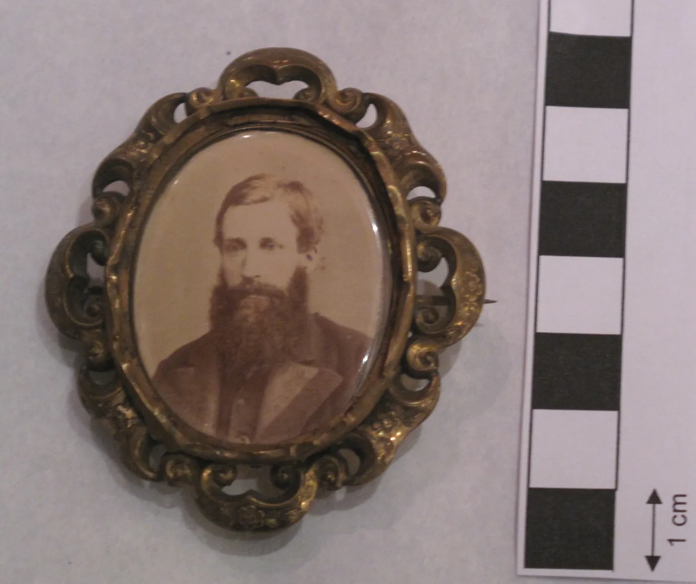 Gilt-metal photographic portrait brooch with a pierced scroll border framing a sepia photograph of a bearded man, a fine pin visible along the right-hand edge, beside a centimetre scale bar