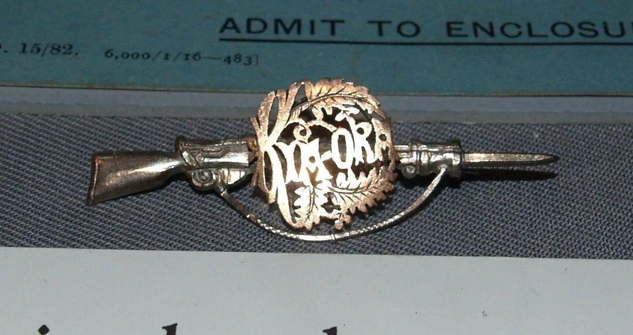 Back of a silver sweetheart brooch shaped as a rifle with a KIA-ORA wreath, fitted with a plain wire C-catch and a long pin whose point projects past it