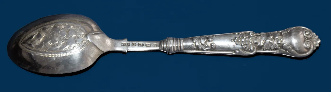 Ornate Victorian silver serving spoon photographed against a deep blue ground, with a row of small struck hallmarks on the flat of the stem