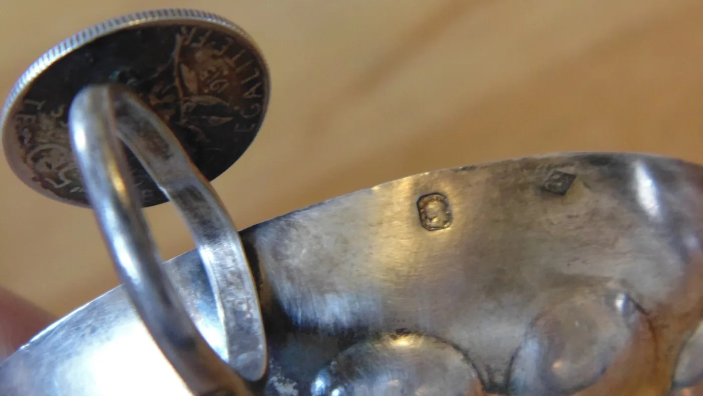 Silver tastevin held up to the light, with small struck punches visible on the curved inner surface near the thumb rest