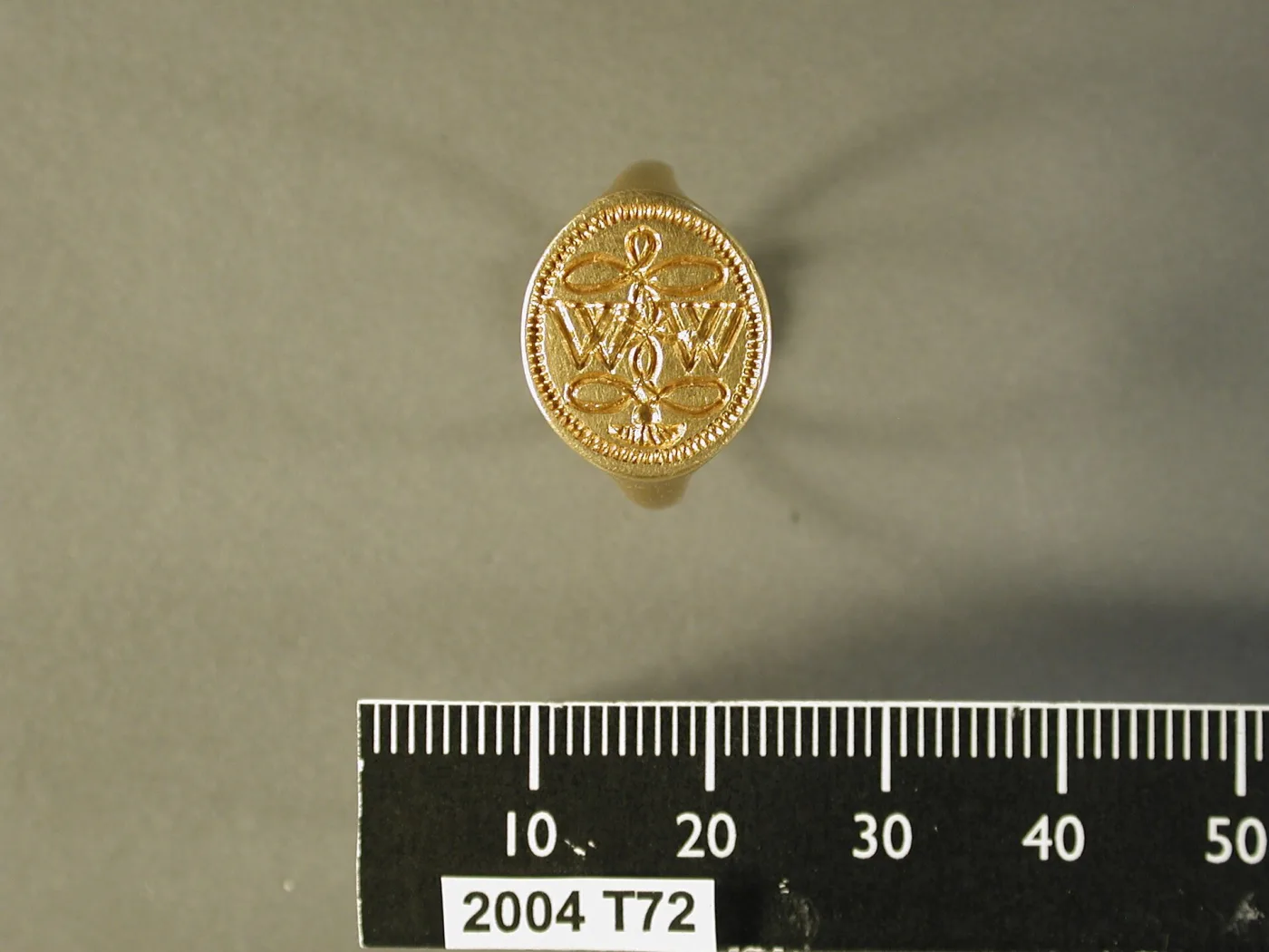 Small gold signet ring lying beside a centimetre scale, the flat oval bezel engraved with a monogram