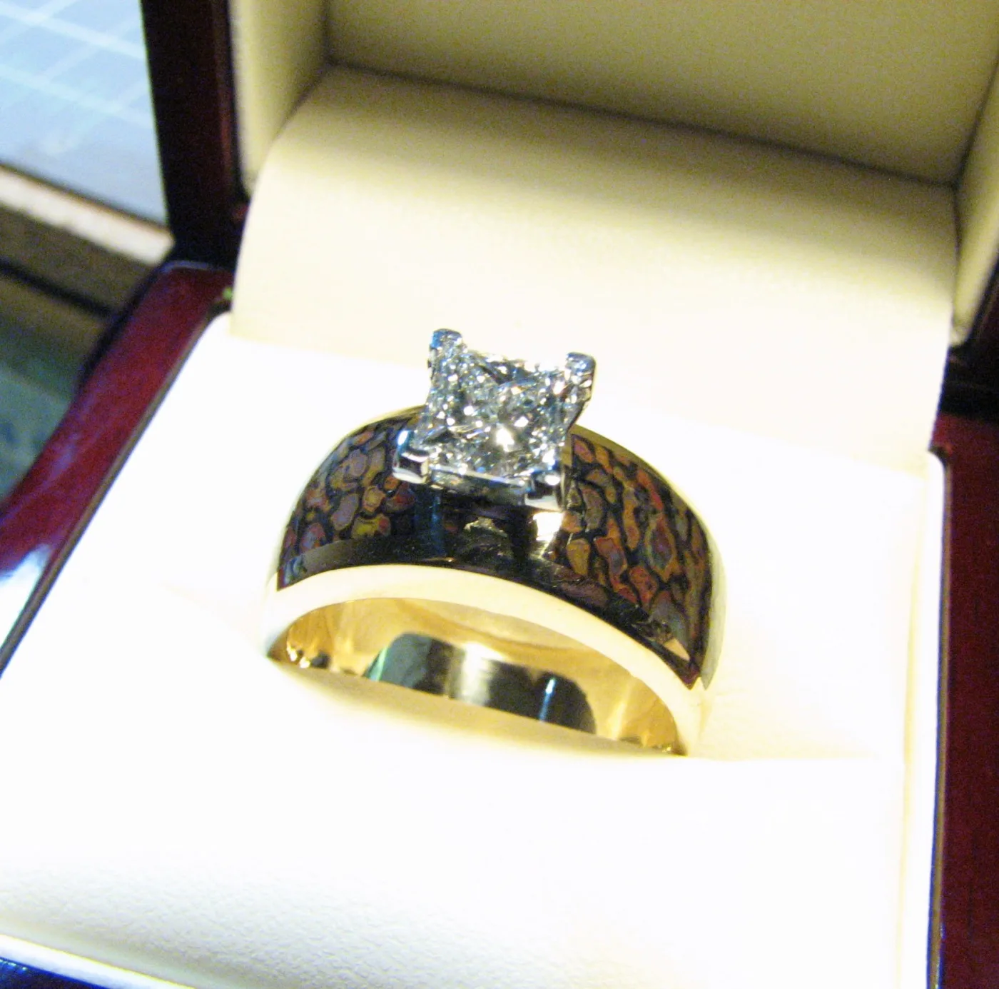 A wide yellow gold wedding ring with a mottled brown inlay around the band and a large square princess-cut diamond in a white metal setting, in a white ring box