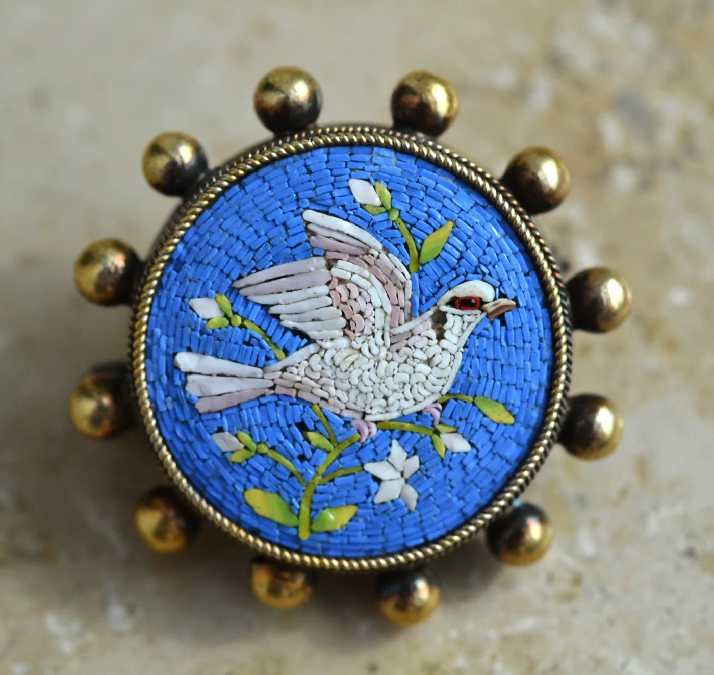Round brooch worked in tiny blue and white glass tesserae showing a white dove with an olive sprig, in a gilt rope-twist bezel ringed with metal balls