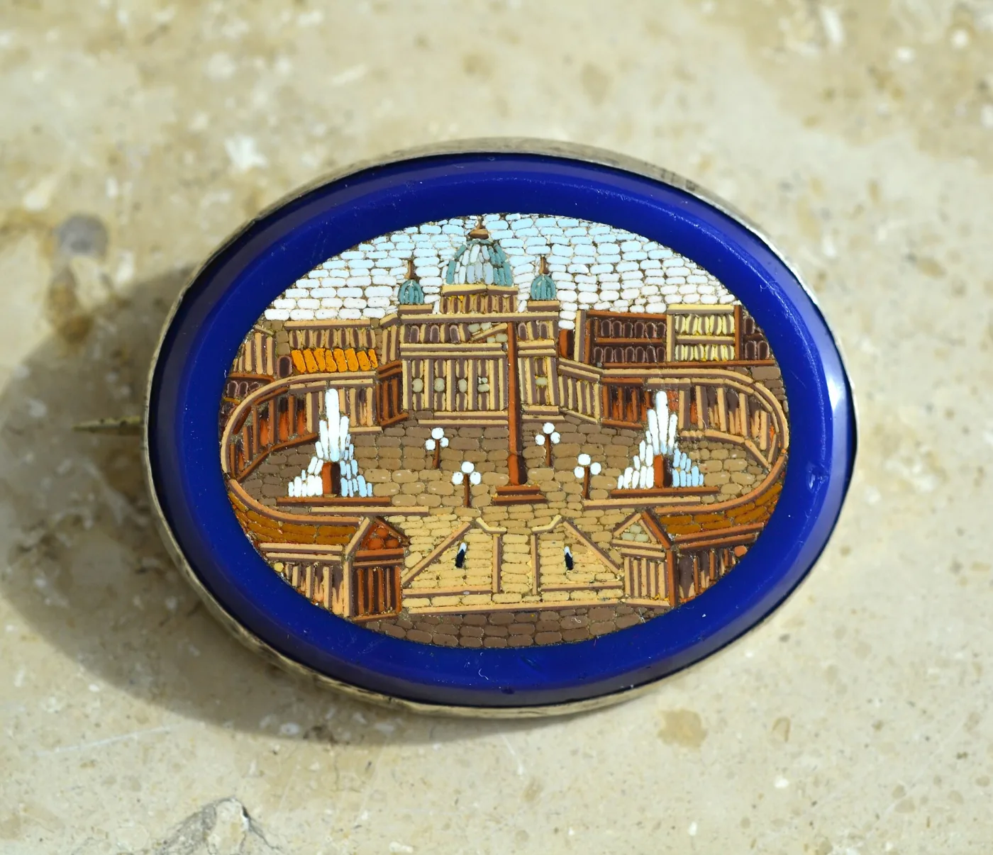 Oval brooch on a cobalt blue glass ground showing Saint Peter's Square with the basilica dome, colonnades and two fountains, the sky visibly made of rows of separate pale oblong tiles