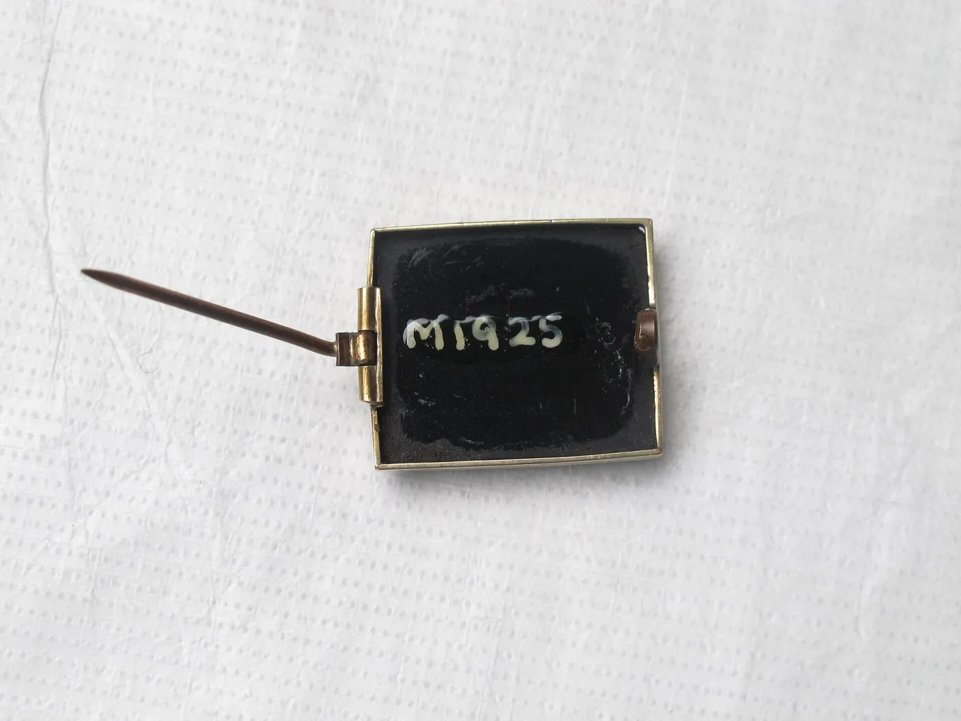 Reverse of the same small rectangular brooch showing a plain black slab in a thin gilt frame, a simple hinged pin extending to the left, and the number M1925 painted in white