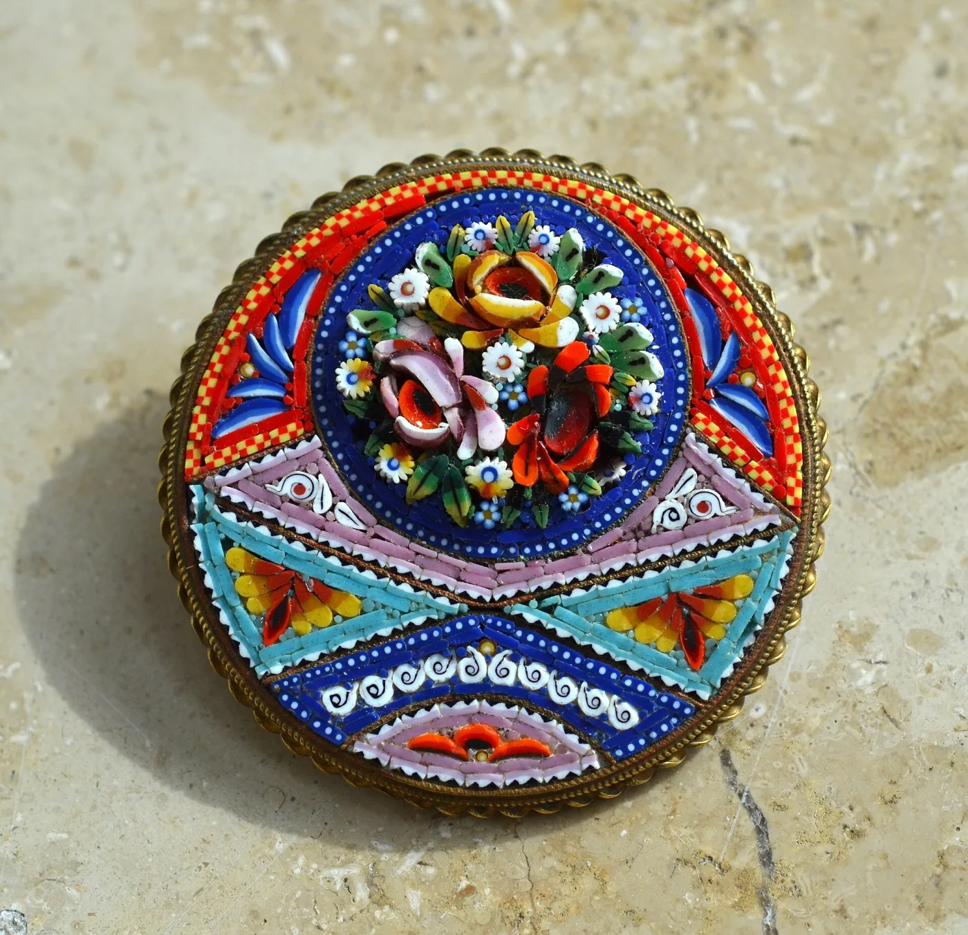 Round brooch in a scalloped gilt brass mount, a central bouquet of glass flowers surrounded by broad bands of red, blue, pink and turquoise geometric mosaic with checkerboard and spiral patterned pieces