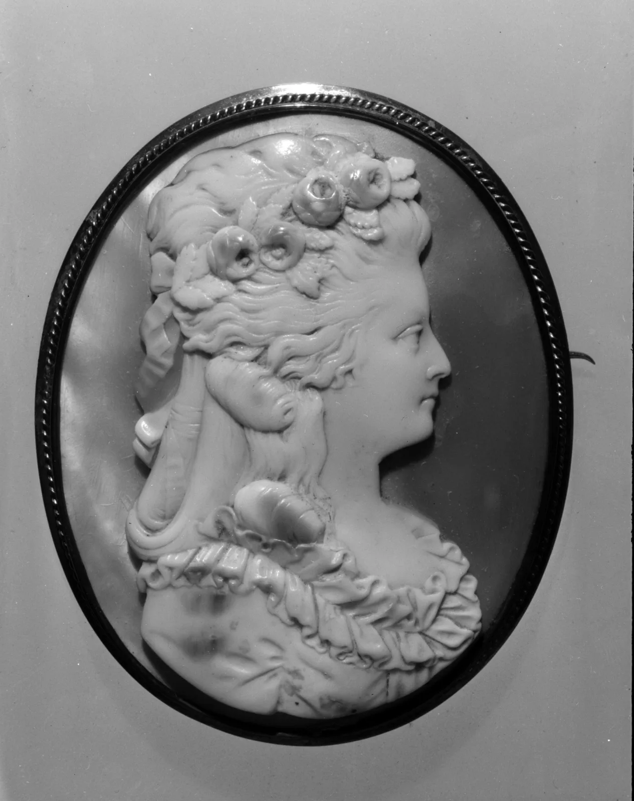 Oval cameo of a woman in right-facing profile with long curled hair dressed with roses and a ruffled 18th-century neckline, in a thin gold frame