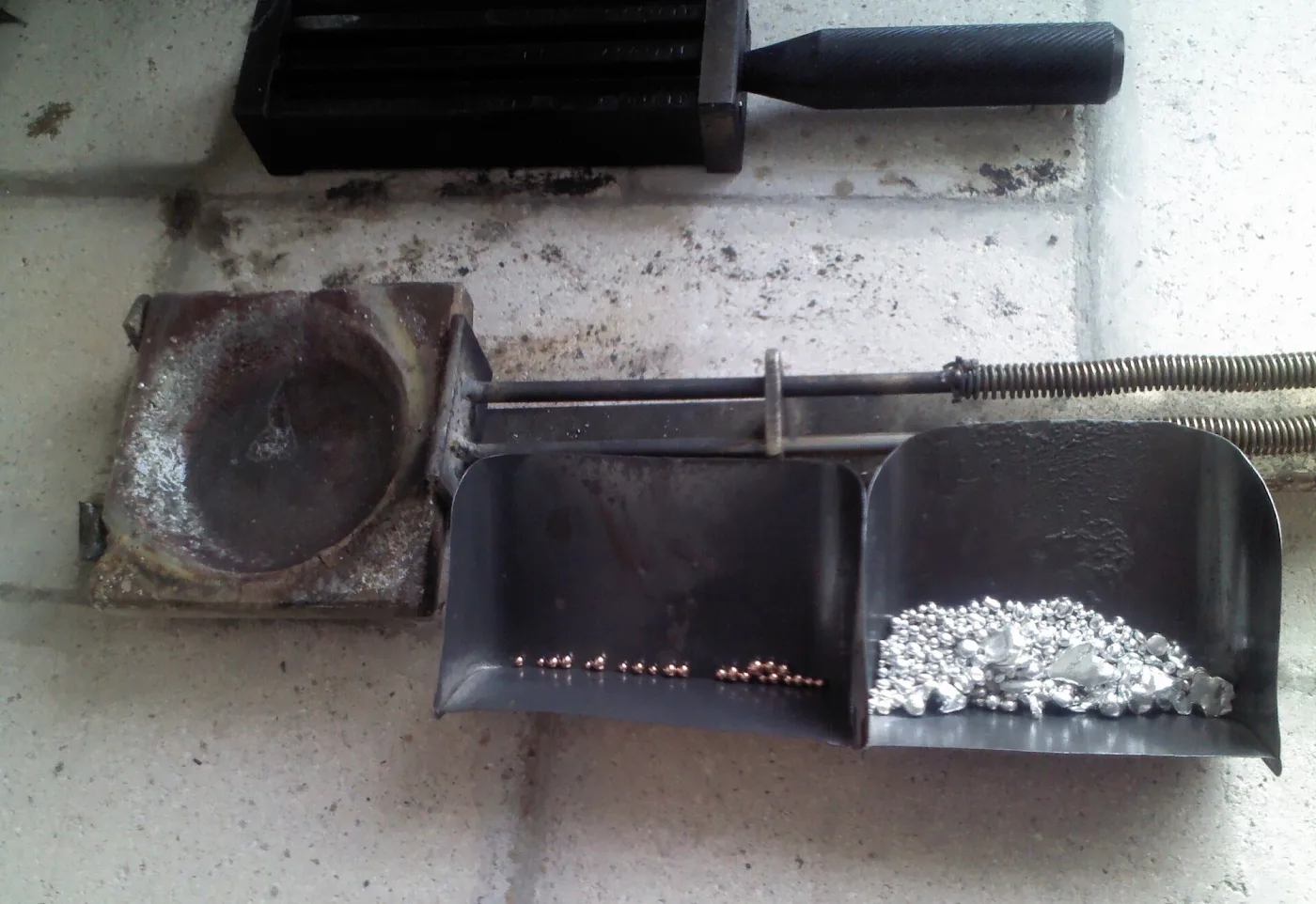 A workbench with a crucible, an ingot mould, and a scoop holding silver granules ready to be melted